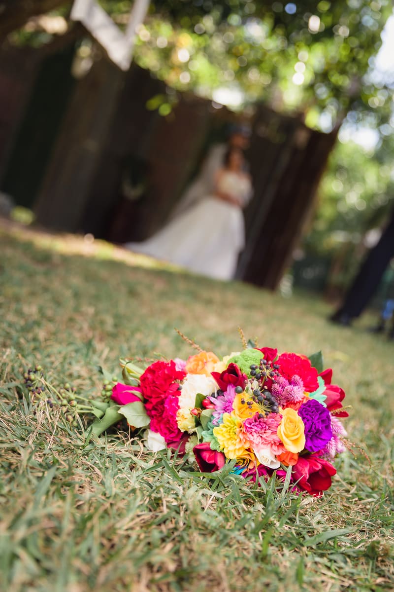 A colorful bridal bouquet lies on the grass with the bride and groom blurred in the background. Wedding industry awards finalist by She Said Yes.