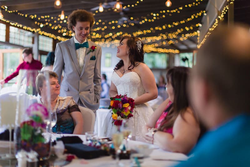 The bride Holly in a white wedding dress holding a colorful bouquet talks with the groom Paul in a light gray suit and teal bow tie at a table with seated guests during the reception at Kwila Lodge.