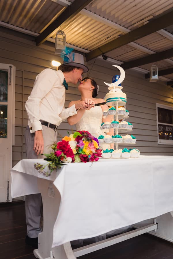 Bride Holly and groom Paul at Kwila Lodge reception stage cutting a tiered cupcake and cake stand decorated with teal and white frosting, with a colorful bridal bouquet on the table.