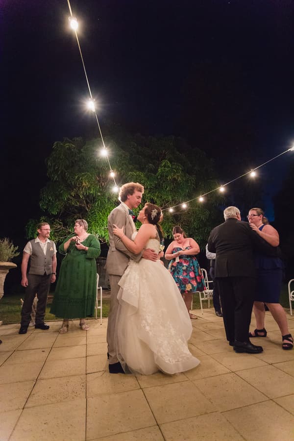 Bride Holly and groom Paul share a dance under string lights at Kwila Lodge while guests dance and mingle nearby.