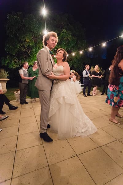 The bride Holly and groom Paul dance together on the outdoor patio, surrounded by guests and string lights overhead. Wedding date changed COVID by She Said Yes.