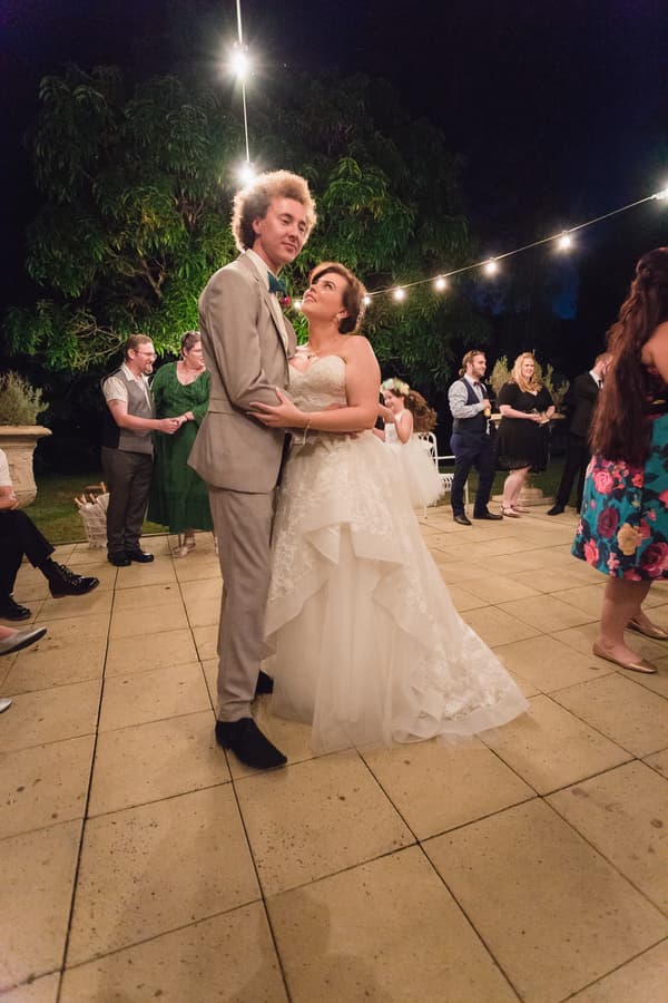 The bride Holly and groom Paul dance together on the outdoor patio at Kwila Lodge during the reception, surrounded by guests and string lights overhead.