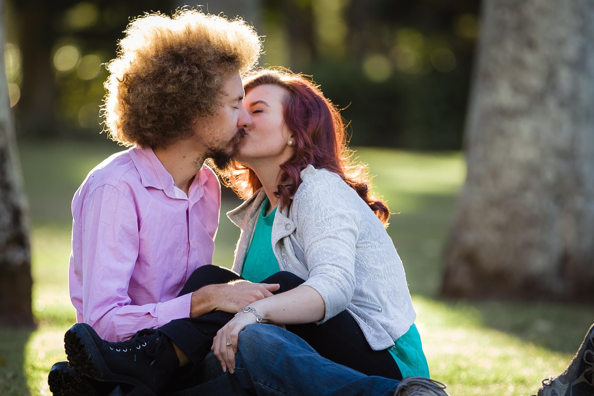 Couple sitting on grass at City Botanic Gardens, Brisbane, sharing a kiss in soft sunlight.