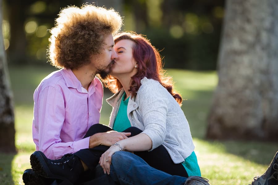 Couple sitting on grass at City Botanic Gardens, Brisbane, sharing a kiss in soft sunlight.