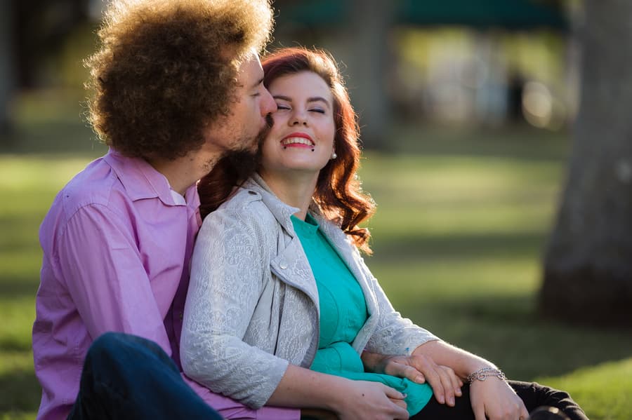 Couple sitting on grass, one kissing the other's cheek, both smiling warmly at City Botanic Gardens.