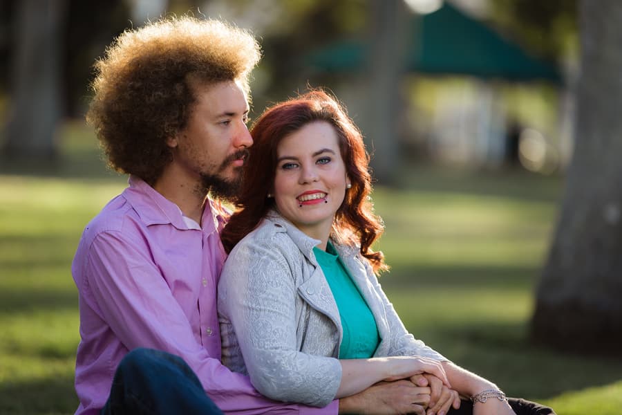 Couple sitting on grass, one looking at the other with a smile, trees in the background.