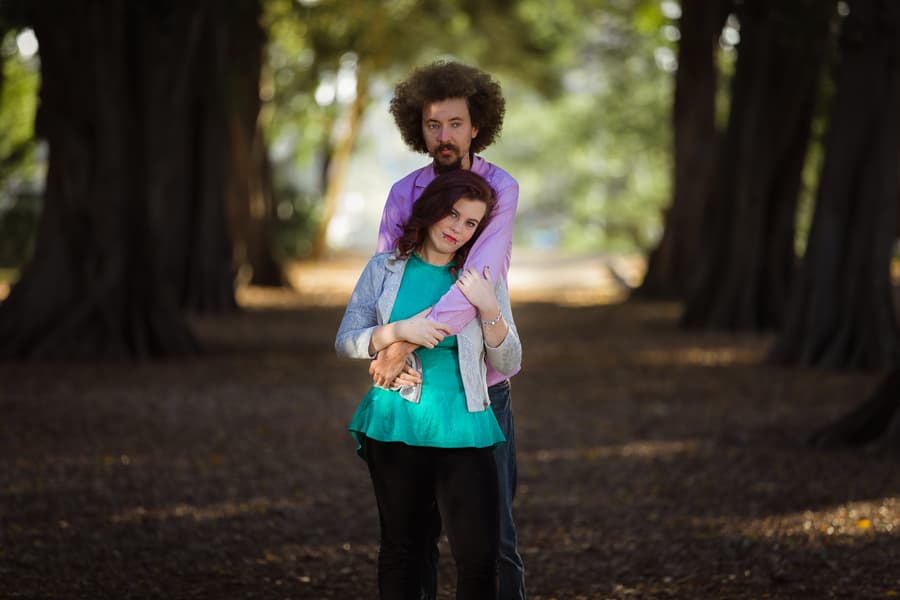 Couple sitting closely on grass, one looking at the other, surrounded by trees.