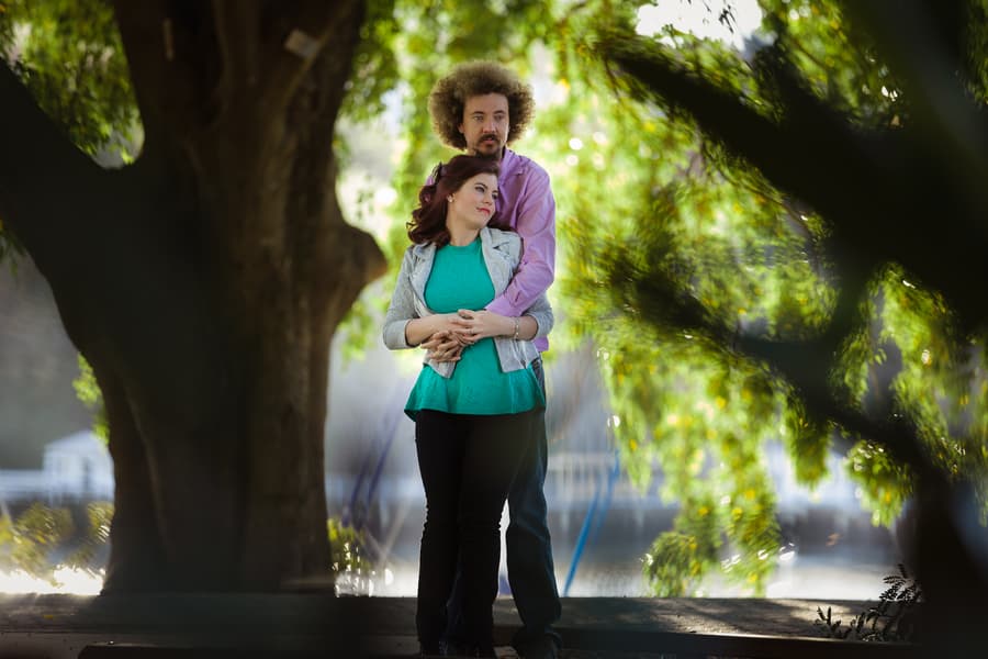 Couple standing close in a tree-lined path, one embracing the other from behind.