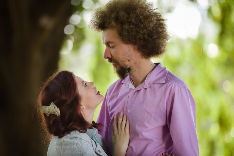 Couple standing under a large tree, one embracing the other from behind, soft light filtering through leaves.