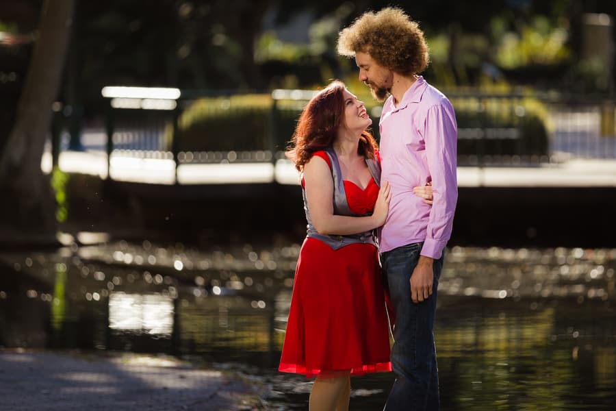 Couple laughing together, standing close with water and trees in the background.