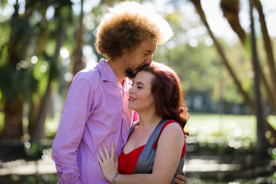 The couple stands closely in City Botanic Gardens, Brisbane, with one kissing the other's forehead under soft sunlight.