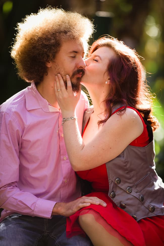 The couple shares a gentle kiss while seated on a stone ledge in City Botanic Gardens, Brisbane, with sunlight filtering through the trees.