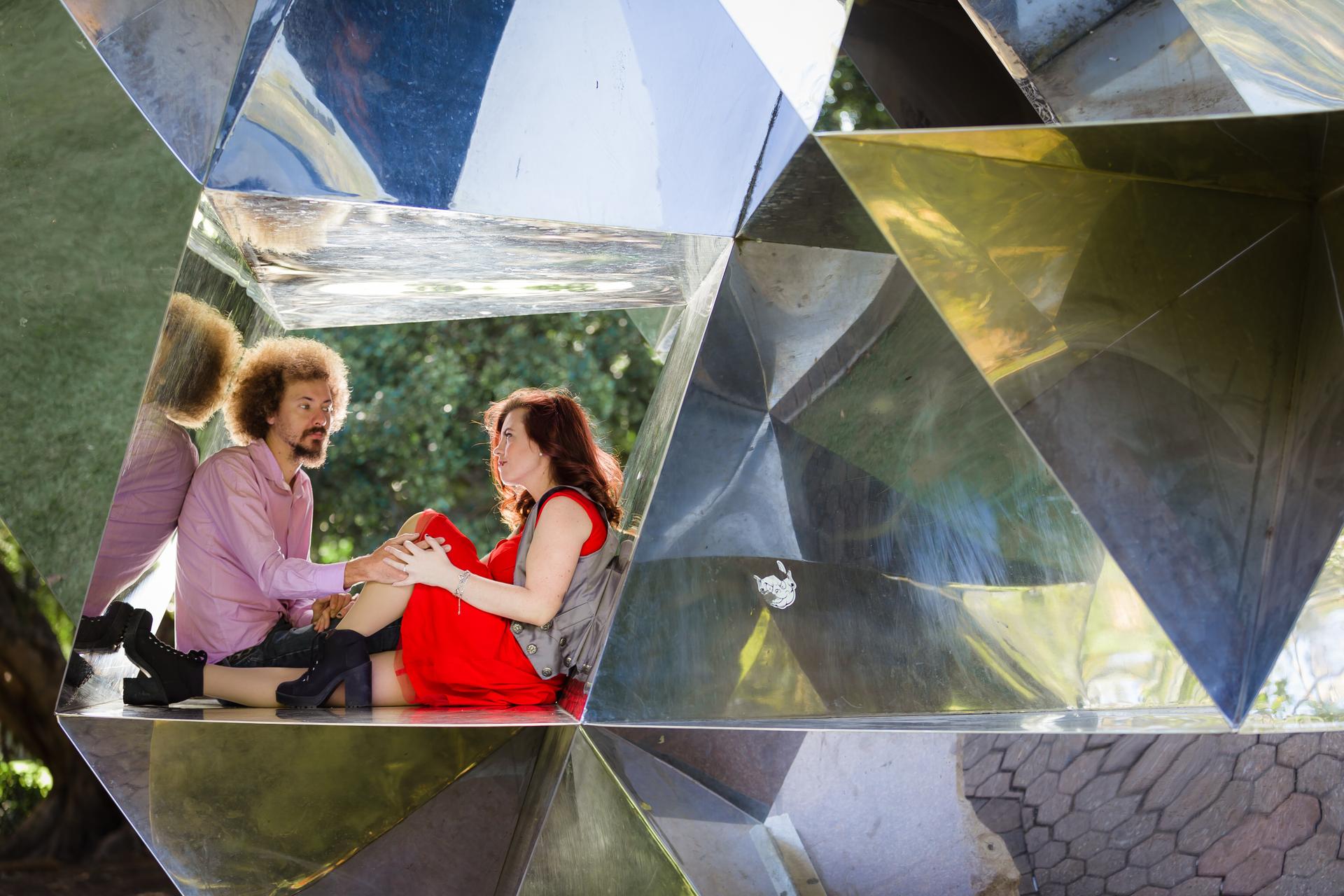 The couple is seated inside a large geometric sculpture in City Botanic Gardens, Brisbane, sharing a joyful interaction.