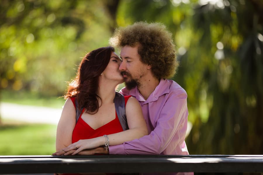 The couple leans on a wooden railing in City Botanic Gardens, Brisbane, sharing a tender kiss with greenery in the background.