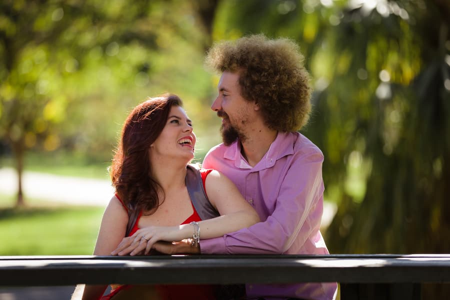 The couple leans on a wooden railing in City Botanic Gardens, Brisbane, smiling warmly at each other with a lush backdrop.