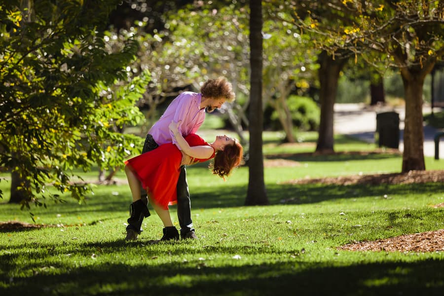 One person dips the other playfully on a grassy area in City Botanic Gardens, Brisbane, surrounded by trees and sunlight.
