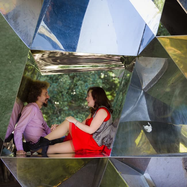 The couple sits inside a reflective geometric sculpture in City Botanic Gardens, Brisbane, engaging in a lively conversation.