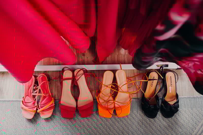 Four pairs of women's shoes in various colors (pink, red, orange, black) arranged on the floor beneath hanging red and patterned dresses.