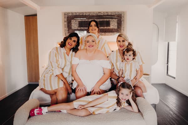 The bride Jacquelyne sits on a bed surrounded by three bridesmaids and two children, all wearing matching striped shirts, in a bright room at Sandstone Point Hotel.