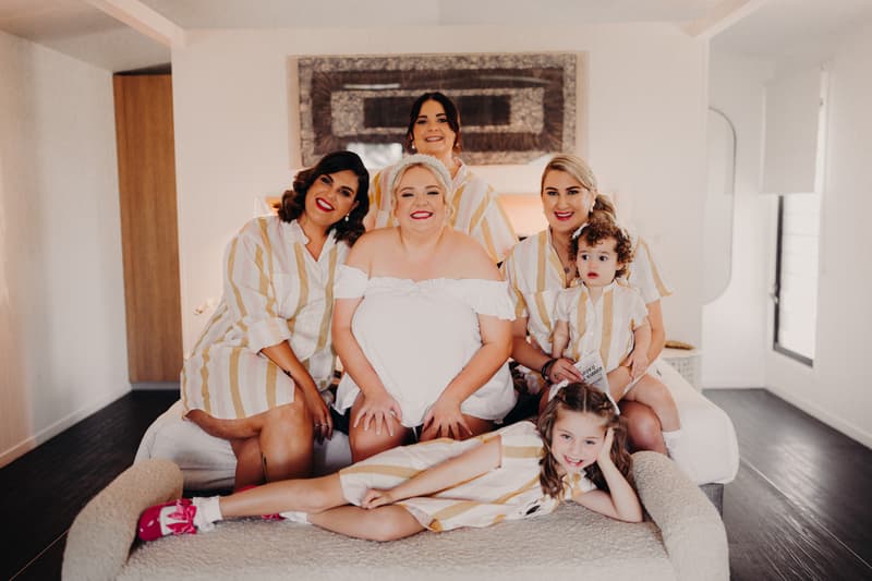 The bride Jacquelyne sits on a bed surrounded by three bridesmaids and two children, all wearing matching striped shirts, in a bright room at Sandstone Point Hotel.