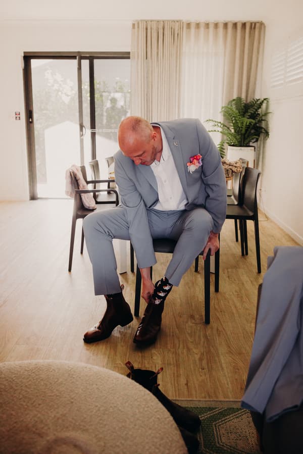 The groom Arran sits on a chair at Sandstone Point Hotel Pavilion, putting on his brown shoes and showing patterned socks.