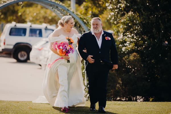 The bride Jacquelyne walks arm-in-arm with an older man, likely her father, outdoors at Sandstone Point Hotel — Pavilion, holding a vibrant bouquet of flowers.