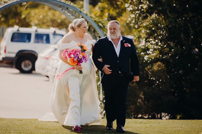 The bride Jacquelyne walks arm-in-arm with an older man, likely her father, outdoors at Sandstone Point Hotel — Pavilion, holding a vibrant bouquet of flowers.