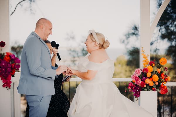 Jacquelyne and Arran exchange rings during their wedding ceremony at Sandstone Point Hotel — Pavilion, with the officiant holding a microphone between them and floral arrangements on either side.
