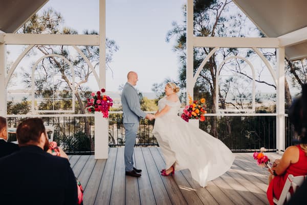 Bride Jacquelyne and groom Arran hold hands facing each other during their wedding ceremony at Sandstone Point Hotel — Pavilion, with guests seated and floral arrangements on white pillars behind them.