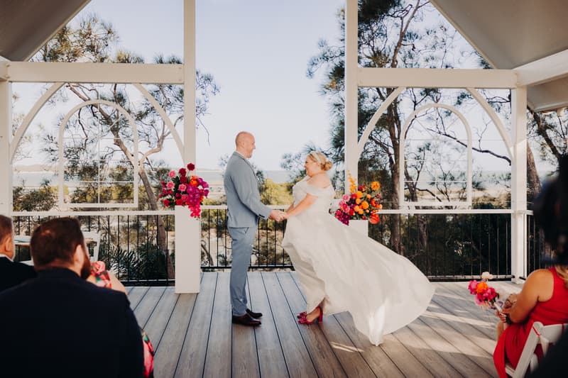 Bride Jacquelyne and groom Arran hold hands facing each other during their wedding ceremony at Sandstone Point Hotel — Pavilion, with guests seated and floral arrangements on white pillars behind them.