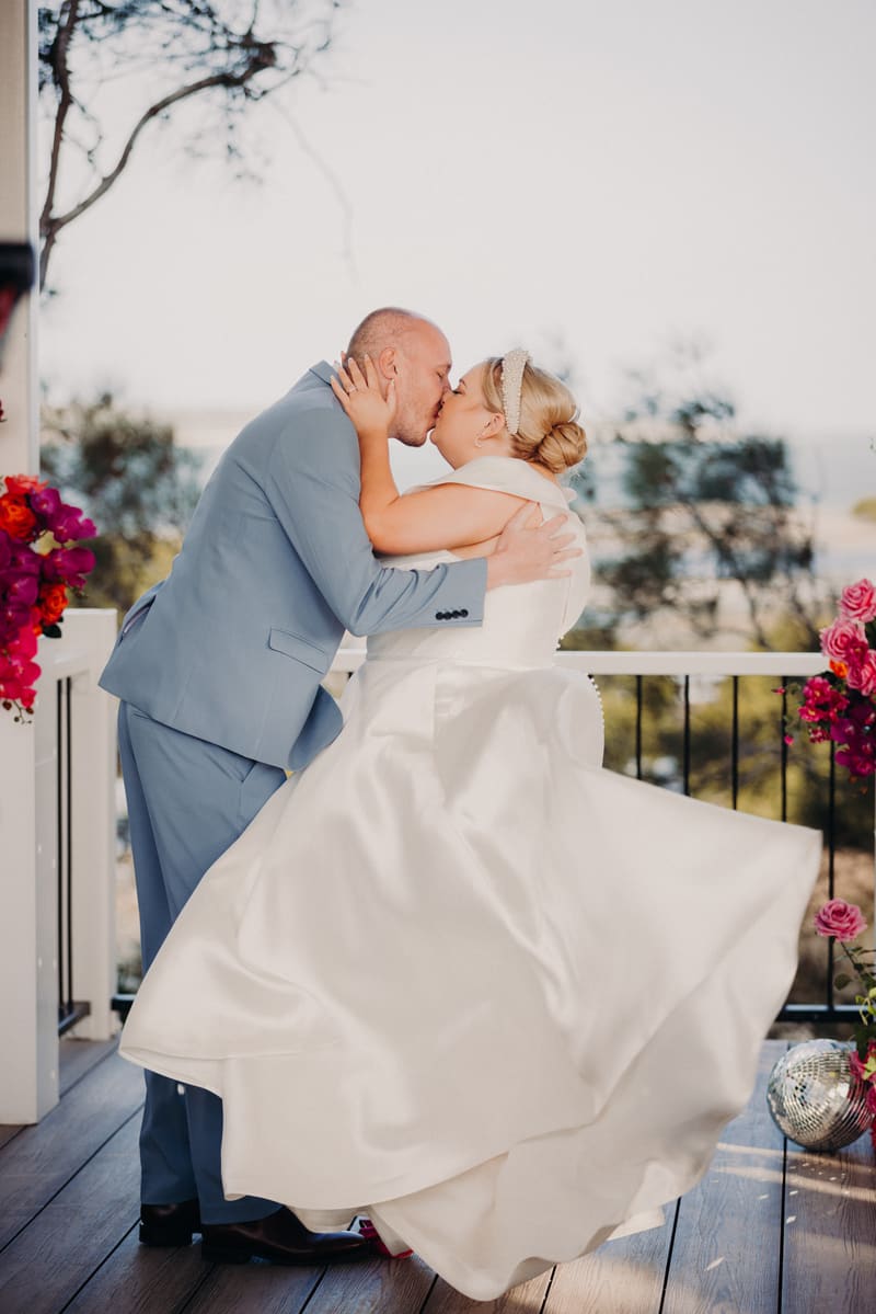 Bride Jacquelyne and groom Arran share a kiss on a balcony, surrounded by floral arrangements. Candid wedding photographer by She Said Yes.