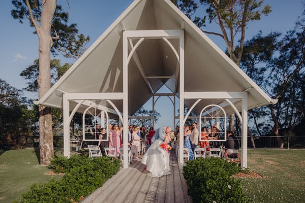 Bride Jacquelyne and groom Arran share a kiss under the pavilion at Sandstone Point Hotel during their wedding ceremony, with guests seated and standing on either side of the aisle.