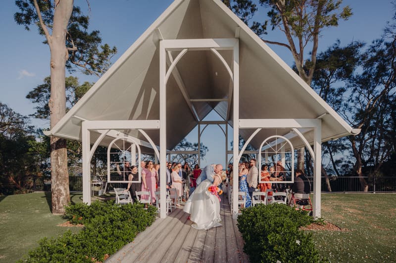 Bride Jacquelyne and groom Arran share a kiss under the pavilion at Sandstone Point Hotel during their wedding ceremony, with guests seated and standing on either side of the aisle.