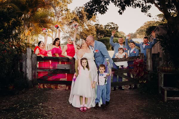 Jacquelyne and Arran kiss in front of a wooden gate at Sandstone Point Hotel, surrounded by bridesmaids in pink dresses and groomsmen in blue suits, with flower girls and a ring bearer in front.
