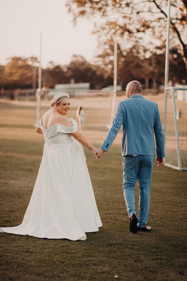 The bride Jacquelyne in a white off-shoulder wedding gown and the groom Arran in a light blue suit hold hands walking on grass near soccer goals at Sandstone Point Hotel.