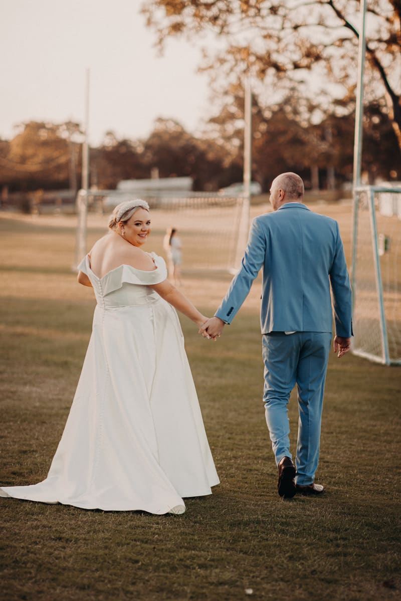 The bride Jacquelyne in a white off-shoulder wedding gown and the groom Arran in a light blue suit hold hands walking on grass near soccer goals. Engagment party by She Said Yes.