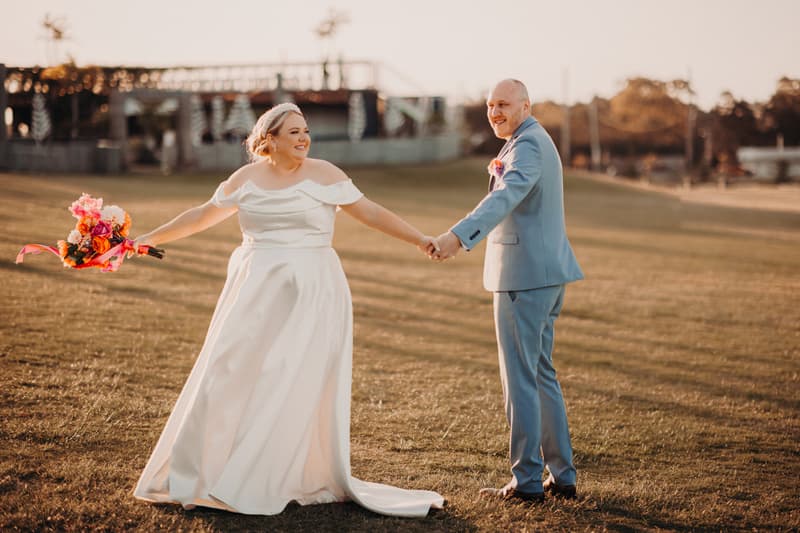 The bride Jacquelyne and groom Arran hold hands outdoors on a grassy field at Sandstone Point Hotel during their couple portraits session. Jacquelyne wears a white off-shoulder wedding gown and holds a colorful bouquet, while Arran wears a light blue suit with a boutonniere.