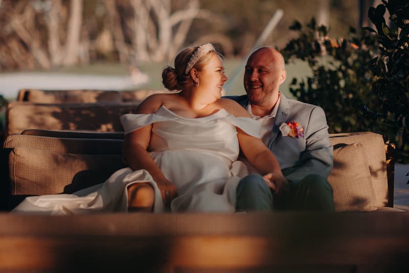The bride Jacquelyne and groom Arran sit closely together on an outdoor couch at Sandstone Point Hotel, sharing a moment while looking at each other.