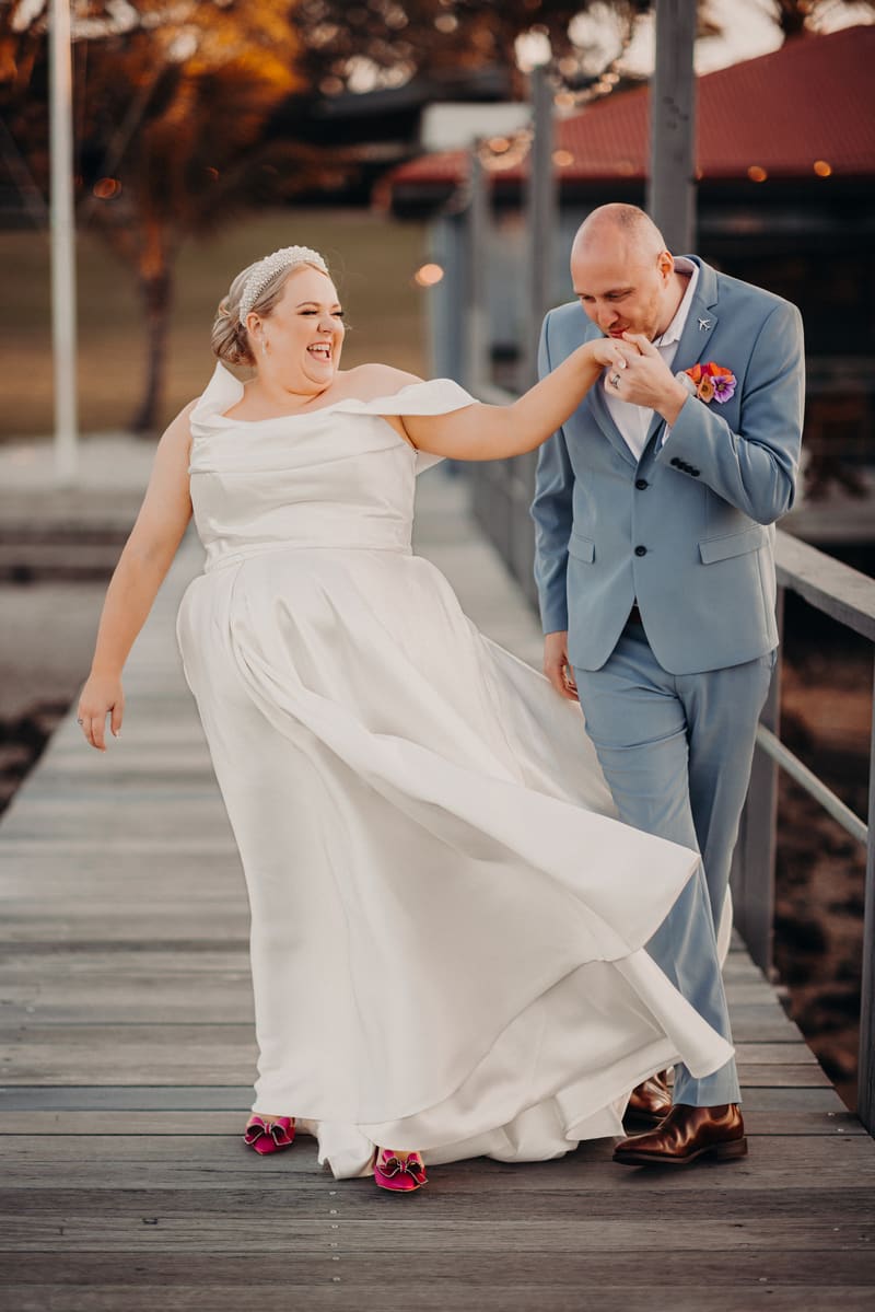 The bride Jacquelyne in a white wedding dress and pink shoes and the groom Arran in a light blue suit with a floral boutonniere pose on a wooden boardwalk at Sandstone Point Hotel. Arran is kissing Jacquelyne's hand while she smiles and lifts her dress slightly.