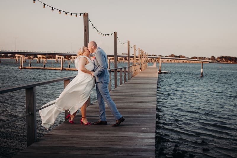 The bride Jacquelyne and groom Arran kiss on a wooden pier at Sandstone Point Hotel with water and a distant bridge in the background during their couple portraits session.