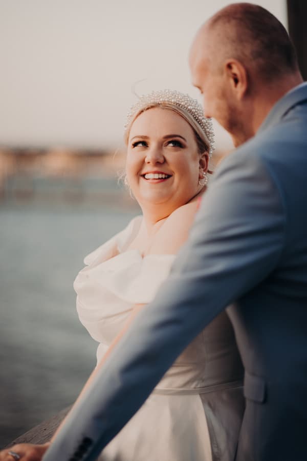 The bride Jacquelyne and groom Arran stand close together by the water at Sandstone Point Hotel during their couple portraits session. Jacquelyne wears a white wedding dress and a pearl headpiece, looking up and smiling at Arran, who is dressed in a light blue suit.