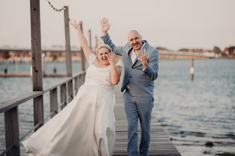 The bride Jacquelyne and groom Arran stand on a wooden jetty at Sandstone Point Hotel, both raising their hands and showing their wedding rings with joyful expressions.