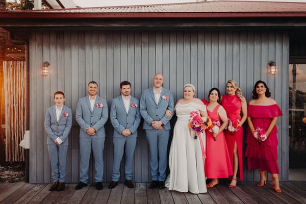 Jacquelyne the bride and Arran the groom stand with their wedding party. The groom and three groomsmen wear light blue suits with boutonnieres, while the bride wears a white gown and holds a colorful bouquet. Four bridesmaids stand beside the bride wearing various shades of pink dresses and holding bouquets. Easter long weekend wedding Brisbane by She Said Yes.