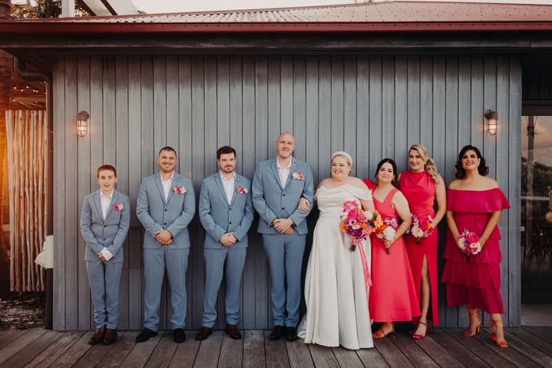 Jacquelyne the bride and Arran the groom stand with their wedding party at Sandstone Point Hotel. The groom and three groomsmen wear light blue suits with boutonnieres, while the bride wears a white gown and holds a colorful bouquet. Four bridesmaids stand beside the bride wearing various shades of pink dresses and holding bouquets.