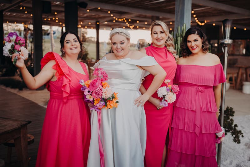 The bride Jacquelyne poses with three bridesmaids in bright pink dresses at Sandstone Point Hotel, each holding bouquets of flowers.