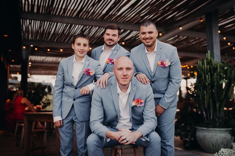 Four men in matching light blue suits with floral boutonnieres pose together under a wooden pergola at Sandstone Point Hotel. One man is seated on a stool while the other three stand behind him, resting their hands on his shoulders.