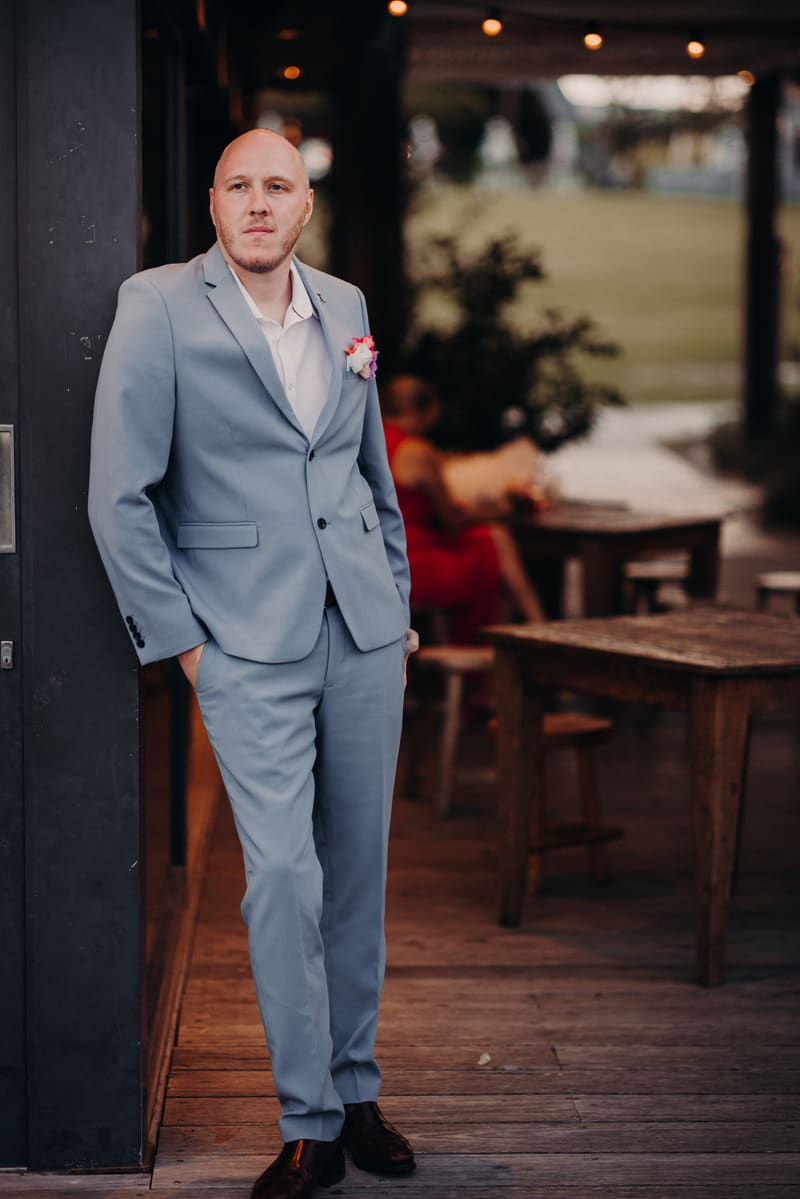 The groom Arran in a light blue suit with a boutonniere leans against a dark pillar at Sandstone Point Hotel, with a blurred figure in a red dress sitting at a wooden table in the background.