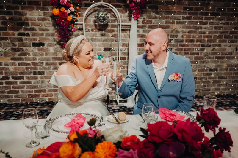 Bride Jacquelyne and groom Arran sit at a reception table, toasting with champagne glasses in front of a brick wall decorated with floral arrangements and a disco ball. Elopement photography packages Queensland by She Said Yes.