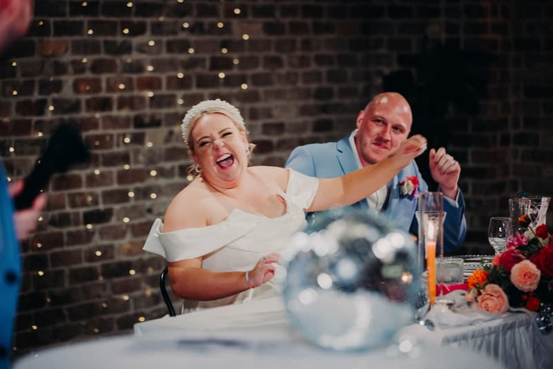 The bride Jacquelyne and groom Arran sit at a table at the Sandstone Point Hotel — Cellar during the reception stage, with Jacquelyne laughing and raising her arm while Arran looks on.