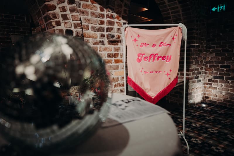 A pink banner with red fringe reading 'Mr & Mrs Jeffrey Est. 02.10.24' is displayed on a stand against a brick wall at Sandstone Point Hotel — Cellar.